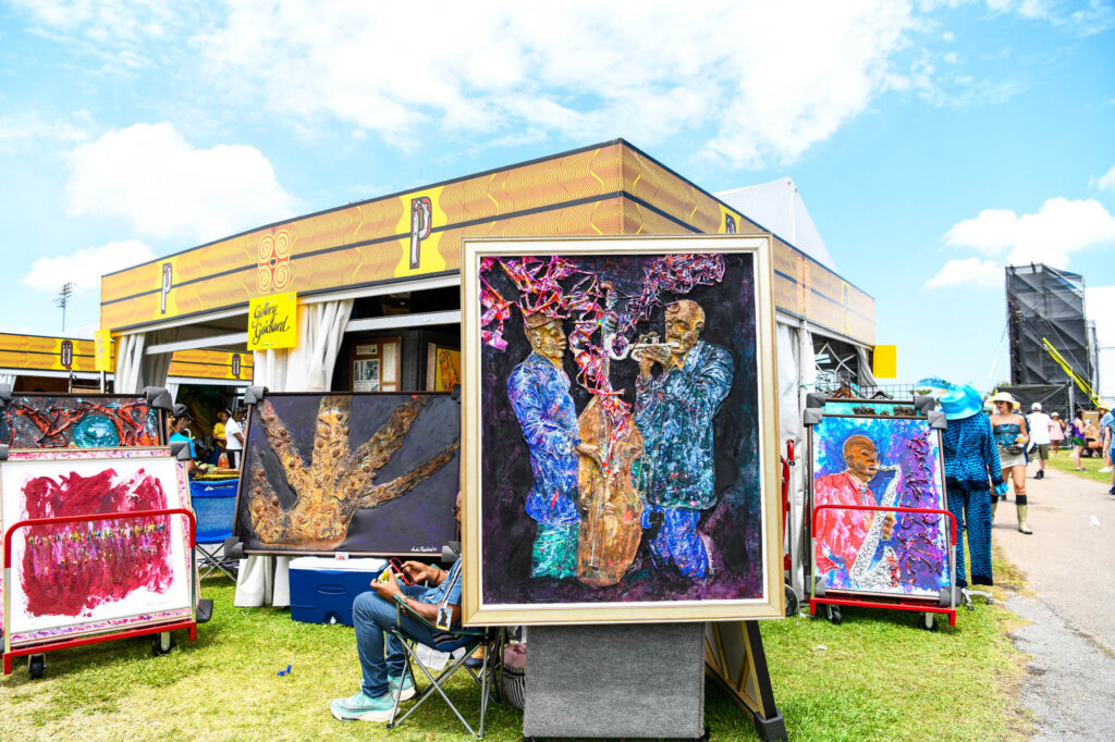 _D850231 - International Magazine Kreol Outdoor art fair scene with colorful, textured paintings on display panels and people strolling among stalls on a sunny day.
