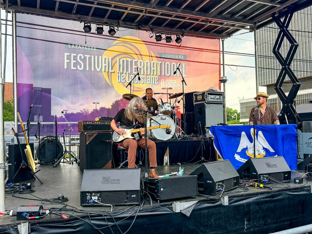 IMG_2850 - International Magazine Kreol Live band on an outdoor stage with guitar, drums, and keyboard; colorful festival backdrop behind them.