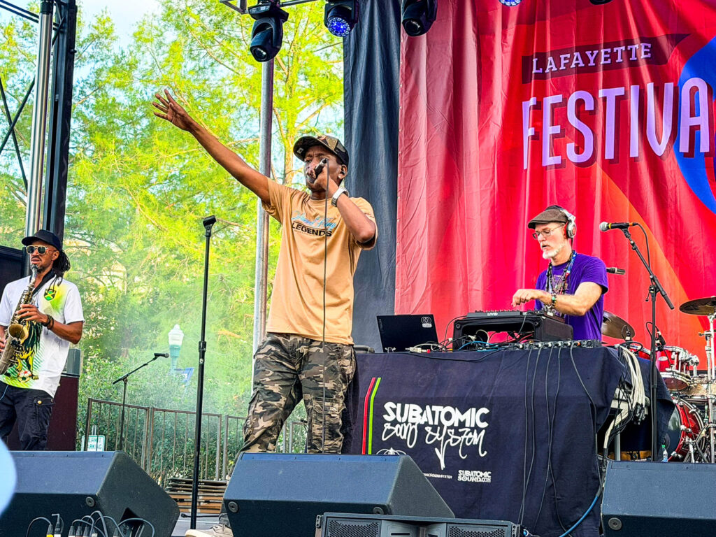 IMG_2912 - International Magazine Kreol Male singer on stage raises his arm and drums into a microphone during a Lafayette Festival performance, with a saxophonist on the left and a DJ on the right behind him.