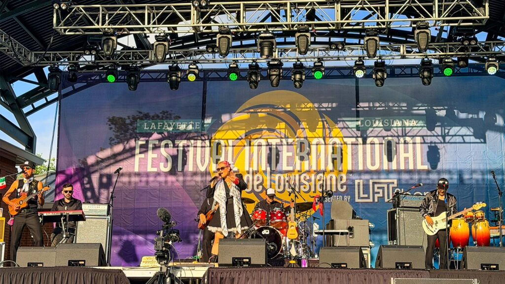 International Festival - International Magazine Kreol Live band performing on an outdoor festival stage with musicians and a large banner reading 'Festival International de Louisiane' in Lafayette, LA.