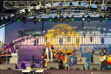 Live band performing on an outdoor festival stage with musicians and a large banner reading 'Festival International de Louisiane' in Lafayette, LA.