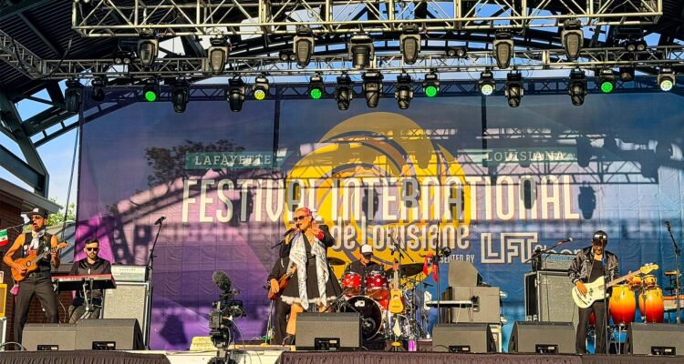 International Festival - International Magazine Kreol Live band performing on an outdoor festival stage with musicians and a large banner reading 'Festival International de Louisiane' in Lafayette, LA.