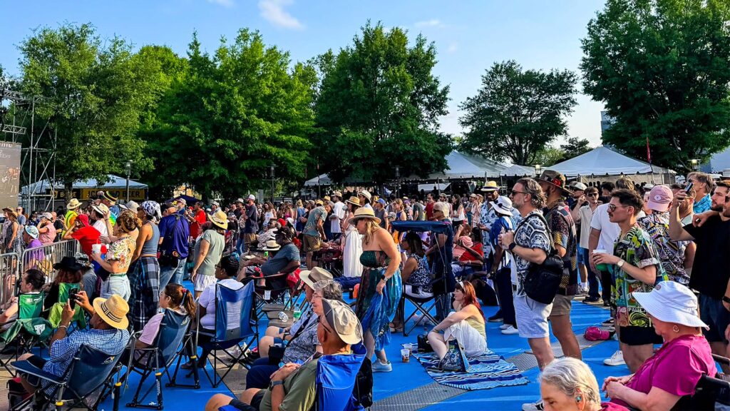 Crowd of people at an outdoor festival or concert, many standing or sitting on a blue platform with tents and trees in the background.