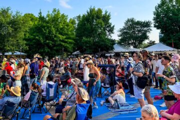 Crowd of people at an outdoor festival or concert, many standing or sitting on a blue platform with tents and trees in the background.