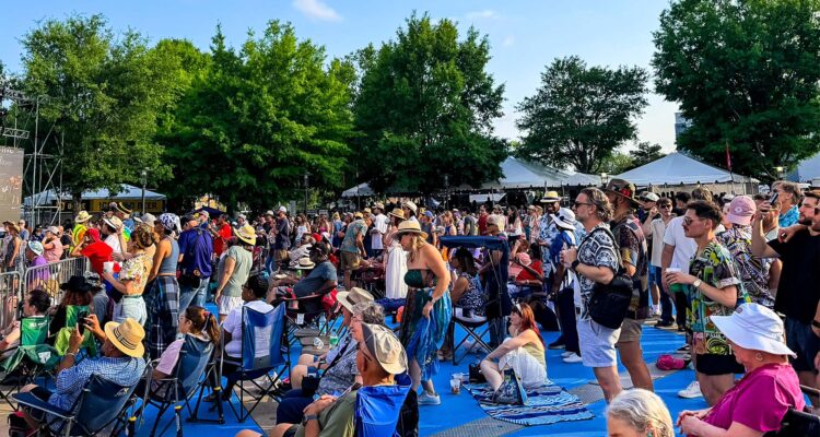 Jazz - International Magazine Kreol Crowd of people at an outdoor festival or concert, many standing or sitting on a blue platform with tents and trees in the background.