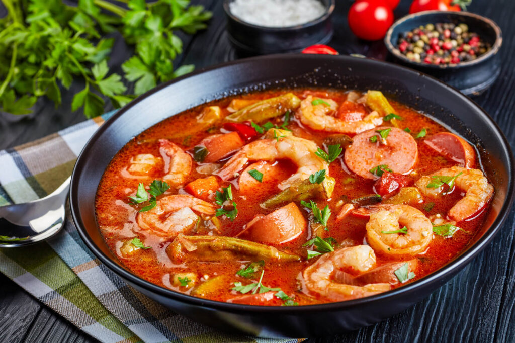 DeliciousGumboWithPrawnsBabyOkraAndSausageInA - International Magazine Kreol Shrimp and sausage gumbo in a dark bowl, garnished with parsley in a red broth.