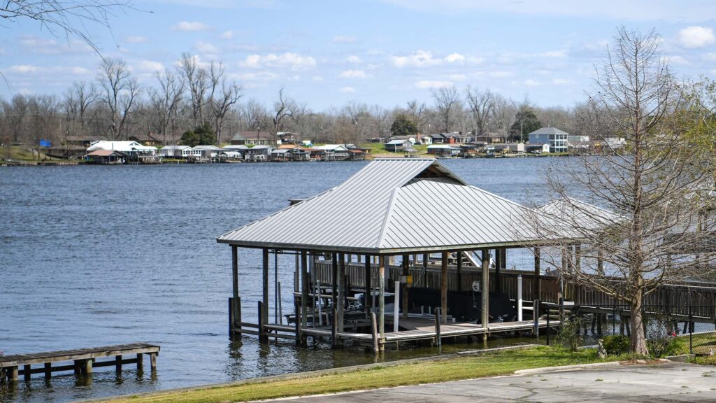 Arnauldville - International Magazine Kreol Boathouse on stilts in a calm lake with a shoreline community across the water and leafless trees under a blue sky
