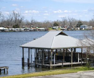 Boathouse on stilts in a calm lake with a shoreline community across the water and leafless trees under a blue sky