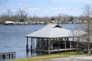 Boathouse on stilts in a calm lake with a shoreline community across the water and leafless trees under a blue sky