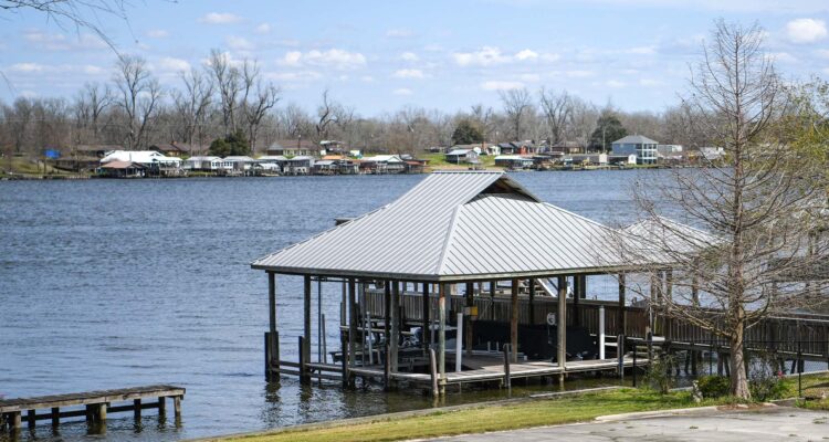 Arnauldville - International Magazine Kreol Boathouse on stilts in a calm lake with a shoreline community across the water and leafless trees under a blue sky