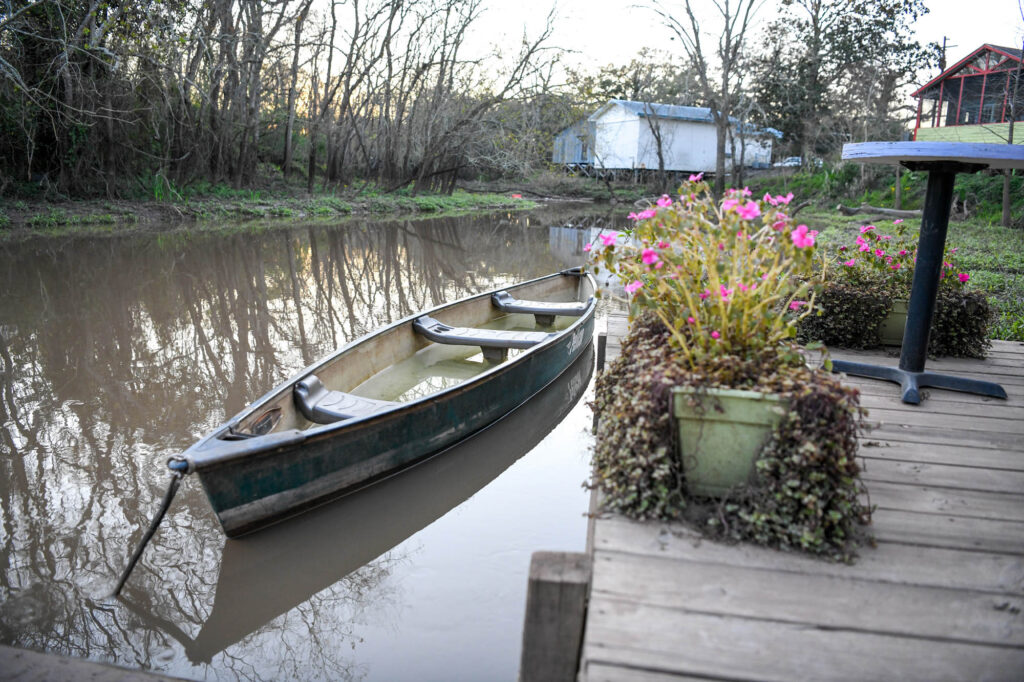 _D851565 - International Magazine Kreol Rowboat tied to a wooden dock along a calm river, pink flowers in the foreground and leafless trees in the background.