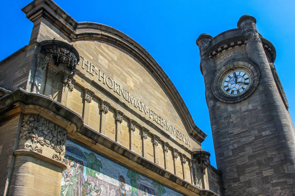 LondonUk-June232020HornimanMuseumClocktower - International Magazine Kreol Historic stone building with a round clock tower on the right against a bright blue sky.
