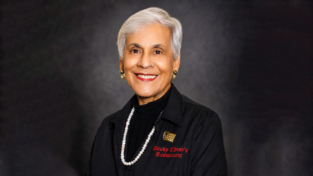 Stella_Lead - International Magazine Kreol Portrait of an elderly woman with short white hair, smiling, wearing a black blazer and pearl necklace against a dark backdrop.