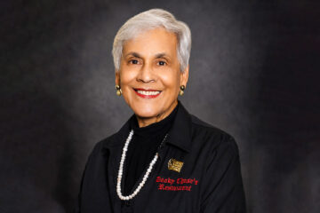 Portrait of an elderly woman with short white hair, smiling, wearing a black blazer and pearl necklace against a dark backdrop.