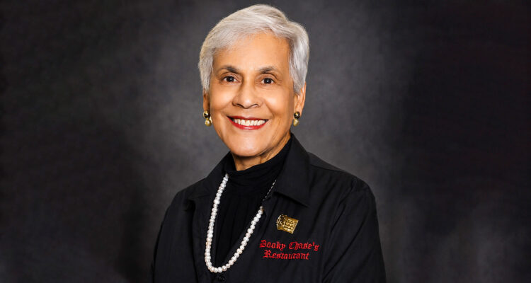 Stella_Lead - International Magazine Kreol Portrait of an elderly woman with short white hair, smiling, wearing a black blazer and pearl necklace against a dark backdrop.