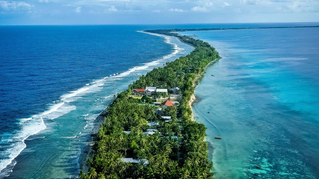 Tuvalu_Lead - International Magazine Kreol Aerial view of a palm-lined isthmus with houses, separating deep blue ocean on the left from a turquoise lagoon on the right.