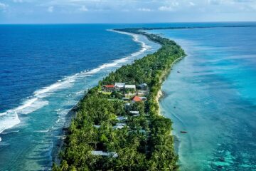 Aerial view of a palm-lined isthmus with houses, separating deep blue ocean on the left from a turquoise lagoon on the right.
