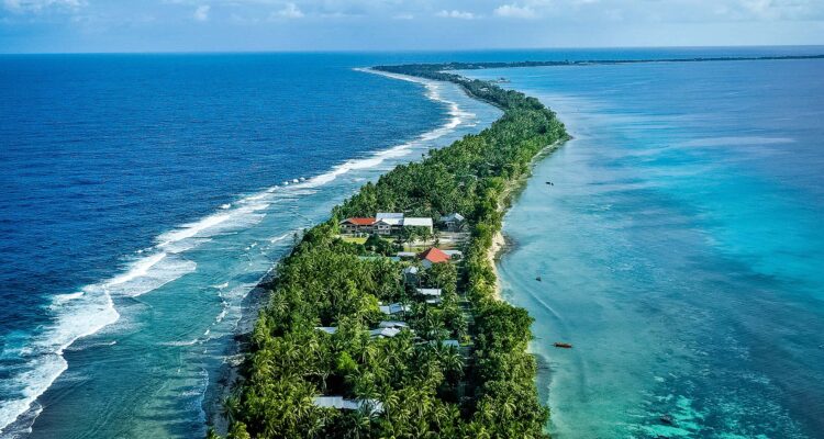 Tuvalu_Lead - International Magazine Kreol Aerial view of a palm-lined isthmus with houses, separating deep blue ocean on the left from a turquoise lagoon on the right.
