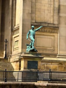 image14 - International Magazine Kreol Bronze-green statue of a soldier in a uniform, arm raised as if signaling, mounted on a stone pedestal in front of a neoclassical building exterior with decorative frieze