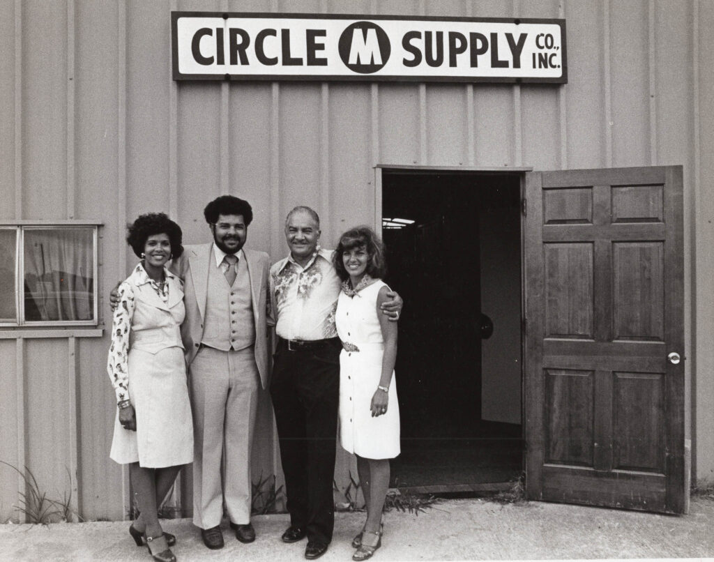 mouton010 - International Magazine Kreol Five adults in 1970s-style clothing pose outside Circle M Supply Co., Inc., in front of a metal building.