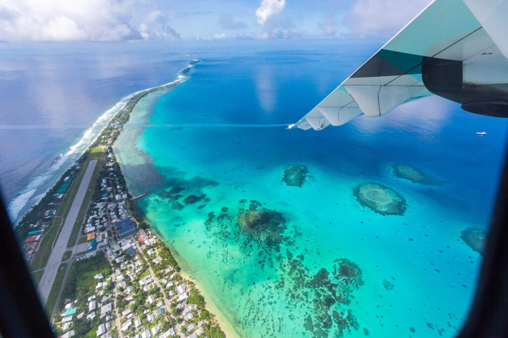 TuvaluIslandLagoonUnderTheWingOfAnAirplaneAerial - International Magazine Kreol Aerial view of a tropical coastline with turquoise reefs, a long island, and a plane wing in the top-right.