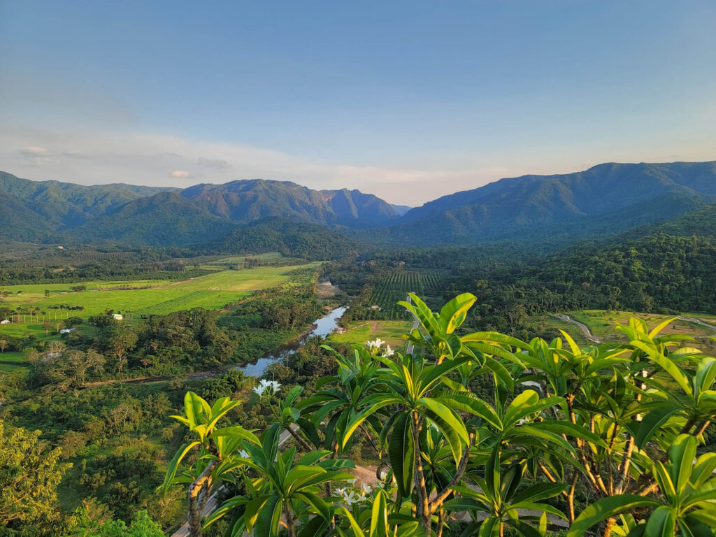SleepingGiantRainforestBelmopanBelize - International Magazine Kreol Valley with green fields and a winding river at the base of forested mountains under a clear blue sky, seen from a foreground viewpoint.