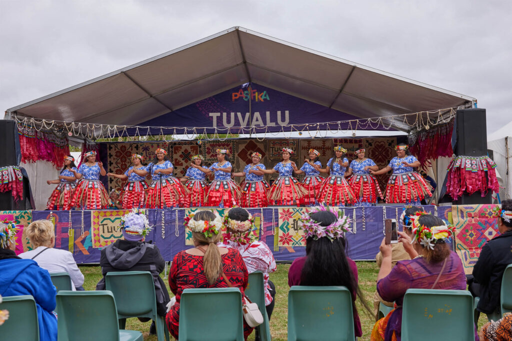 AucklandNewZealand-March9th2024TuvaluTraditionalDance - International Magazine Kreol Group of dancers in blue and white outfits with flower crowns perform on a decorated festival stage, audience seated and watching outdoors.