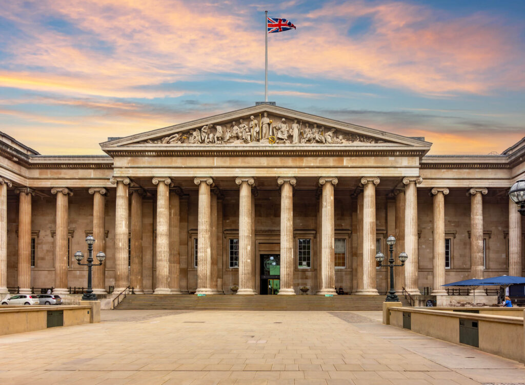 MainEntranceOfBritishMuseumInLondonUk - International Magazine Kreol Front of a neoclassical government building with tall columns, a sculpted pediment, and a flag flying on top at sunset.